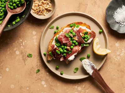 A pea, prosciutto, and ricotta crostini with a bite missing on a plate next to a bowl of peas, walnuts, and flakey sea salt.